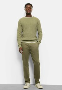 Hellolivgrüner, strukturierter Pullover mit weißem Kragen, kombiniert mit olivgrünen Hosen und cremefarbenen Turnschuhen. Vorderansicht, stehende Haltung.