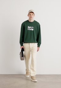 Zelená mikina s bielym logom "Calvin Klein", béžové nohavice a tenisky. Model drží šedú tašku. Jednoduchý dizajn a voľný strih.