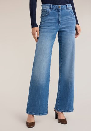 Kvinde iført lyseblå jeans med vidde i benene og knap- og lynlåslukning, kombineret med brune sko med blokhæl og en mørk top med lange ærmer.