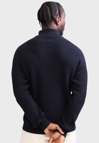 Granatowy sweter z ribbowanej dzianiny z wysokim kołnierzem, długimi rękawami i teksturowymi detalami na rękawach. Widok z tyłu ukazuje dopasowany pas.