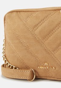 Tan quiltad faux suede axelväska med guldkedjehårdvara, med en logotyp och kontrasterande sömmar. Kompakt och rektangulär form.
