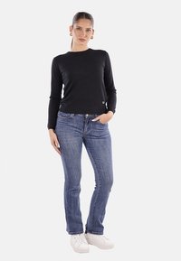 Maglietta nera a maniche lunghe con design aderente, indossata con jeans blu a gamba dritta e sneakers bianche, in piedi contro uno sfondo neutro.