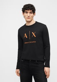 Νέος άνδρας με κατσαρά μαλλιά, ντυμένος με μαύρο μακρυμάνικο πουκάμισο Armani Exchange με πορτοκαλί λογότυπο και μαύρο τζιν, στέκεται μπροστά σε λευκό φόντο.