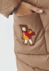 Bruine pufferjas van een gestructureerde stof met een berenpatch op een rood-gele gestreepte trui, geplaatst op de linkerzak.