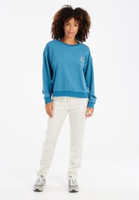 Modrá mikina s okrúhlym výstrihom a dlhými rukávmi, s malým grafickým dizajnom. Skombinovaná s svetlo béžovými nohavicami a sivými teniskami.