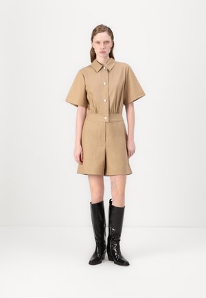 Beige jumpsuit med kort erme, krage, knappelukking foran, elastisk midje og utvidede shorts, kombinert med svarte, glinsende knelengde støvler.