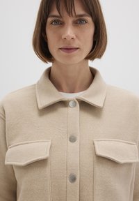 Beige fleece jacket with a collar, two front pockets, and silver snap buttons. The fabric has a soft texture and clean, minimalist lines.