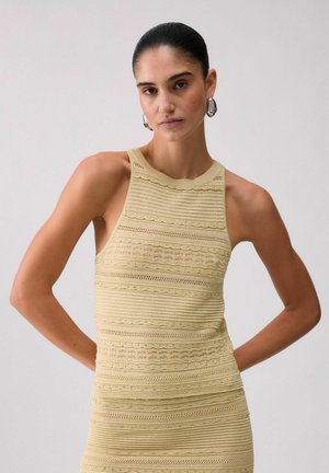 Donna con capelli pettinati all'indietro, che indossa grandi orecchini a cerchio d'argento e un top lavorato a maglia senza maniche color beige con motivi testurizzati.