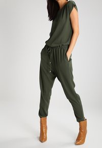 Ensemble deux pièces vert olive comprenant un haut ample avec des manches courtes et un pantalon tapered à taille élastique, associé à des bottes à cheville beiges.