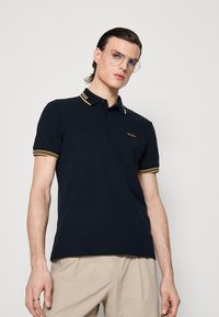 Polo marine avec col et poignets à rayures jaunes. Comprend une patte de boutonnage à trois boutons et un logo discret sur la poitrine. Matière en coton.