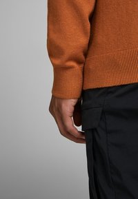 Braccio e mano destra di una persona che indossa un maglione a maglia color ruggine e pantaloni neri con una tasca laterale su uno sfondo grigio.