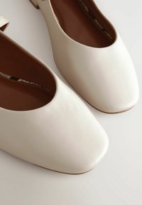 Chaussures plates blanches avec bouts carrés arrondis et semelles intérieures marron sur un fond clair.