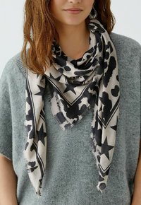 Femme portant un pull en maille gris et une écharpe beige avec des motifs abstraits et des étoiles noires, drapée lâchement autour de son cou.