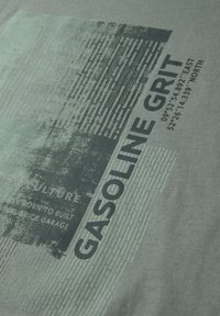 Impression usée avec le texte "GASOLINE GRIT", motifs linéaires abstraits et coordonnées sur un fond de tissu vert pâle.