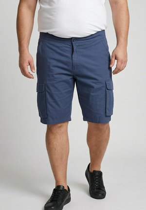 Mann trägt marineblaue Cargo-Shorts mit Klappentaschen, ein weißes Hemd und schwarze Sneakers, steht vor einfarbigem Hintergrund.