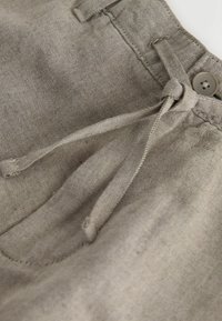 Gros plan de la ceinture d'un pantalon beige avec bouton et cordon en tissu noué, montrant la texture du tissu et les détails des coutures.