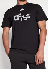 Svart t-shirt gjord av mjuk väv, med en centrerad vit grafisk logotyp och en liten vit logotyp på halsringningen. Korta ärmar.