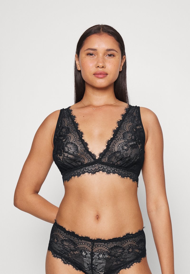 Czarny koronkowy biustonosz w głębokim dekolcie w kształcie V i falbaniastych brzegach, zdobiony kwiatowymi wzorami. W komplecie z pasującymi koronkowymi majtkami, oba bez wyściółki.