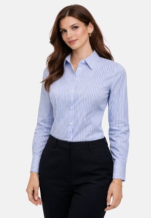 Femme aux longs cheveux bruns portant une chemise à boutons rayée bleue et blanche et un pantalon noir, debout avec une posture détendue sur un fond blanc.