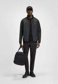 Svart jacka med kontrasterande mörka ärmar, tillsammans med en svart ryggsäck, svarta byxor och svarta skor. Modellen bär en matchande keps.