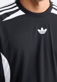 Musta urheilupaita, jossa pystysuorat ohuet raidat, valkoiset korostukset hartioissa ja pieni valkoinen logo keskellä.