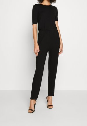 Czarny kombinezon z krótkimi rękawami, elastycznym pasem, prostymi nogawkami i gładką fakturą. W połączeniu z czarnymi sandałami na obcasie.