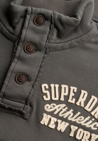 Close-up van een donkergrijze hoodie met drie bruine knopen en geborduurde tekst "Superdry Athletic New York" in crèmewitte draad.