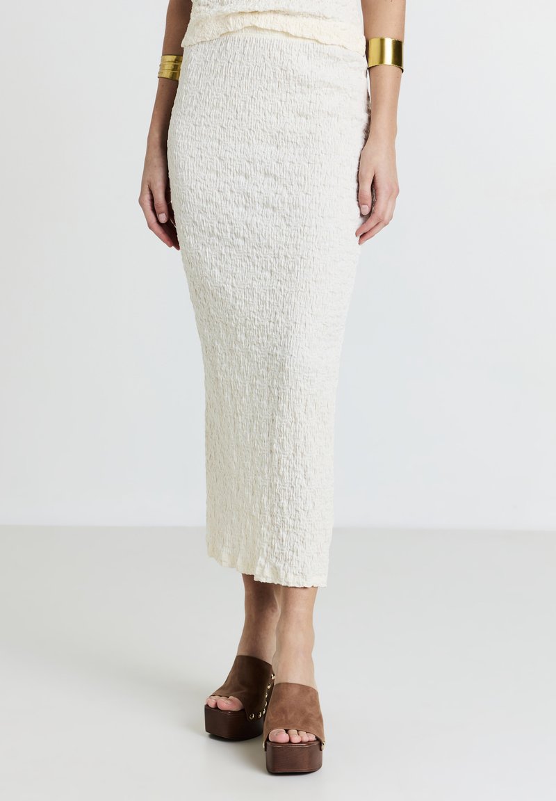 Jupe midi blanche texturée avec un design froissé, assortie à des sandales compensées marron à large sangle et accents dorés.
