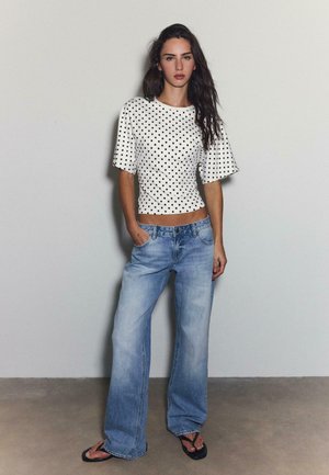 Frau mit langen dunklen Haaren, die ein weißes Kurzarmoberteil mit schwarzen Punkten, ausgewaschene, weit geschnittene Jeans und schwarze Flip-Flops trägt.