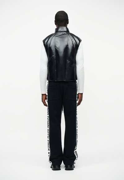 Άτομο που στέκεται με την πλάτη, φοράει ένα γυαλιστερό μαύρο αμάνικο γιλέκο, λευκό πουκάμισο με μακριά μανίκια, μαύρο παντελόνι με λευκές λωρίδες κειμένου και μαύρα παπούτσια.