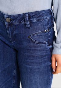 Jeans de mezclilla azul oscuro con cierre de botón, presillas para cinturón y un bolsillo con acentos metálicos, mostrando un detalle de tela texturizada.
