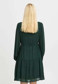 Tummanvihreä mekko, jossa on teksturoitu, raidallinen kuosi. Ominaisuuksia ovat pitkät hihat, kapea vyötärö ja kerroksellinen hame. Näytetään takanäkymä.