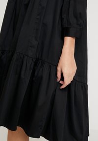 Schwarzes Baumwollkleid mit einem gestuften Design, gepufften Ärmeln und ohne sichtbare Verschlüsse. Glatte Textur mit fließender Silhouette.
