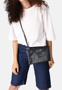 Sac bandoulière en cuir noir avec une surface texturée, doté d'une fermeture éclair et d'un accent en motif tissé sur le devant. Sangle réglable.