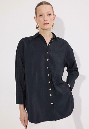 Femeie cu părul tras înapoi, purtând o cămașă neagră lejeră cu nasturi și cercei aurii pendulați, stând cu o mână în buzunar, pe un fundal alb.