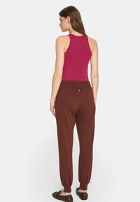 Dopasowany różowy top na ramiączkach i brązowe dresowe spodnie z elastycznymi mankietami oraz pasem. Gładka faktura materiału o podstawowym wzorze. Brązowe obuwie.