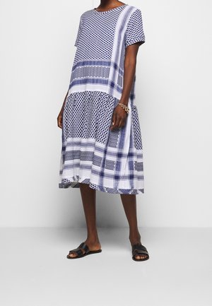 Kobieta w luźnej, niebiesko-białej sukience do kolan w wzory, czarnych, płaskich sandałach i złotej bransoletce, stojąca na tle jednolitego tła.