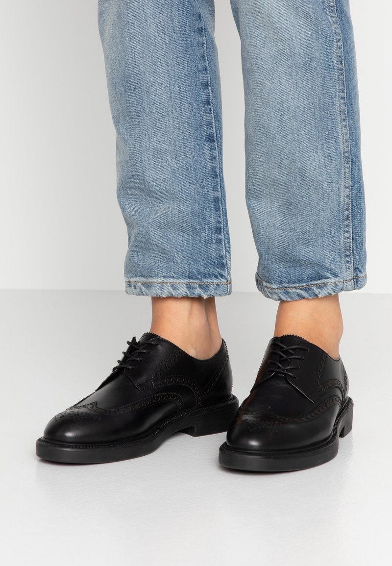 Chaussures habillées en cuir noir avec des perforations brogue détaillées, semelle plate et design traditionnel à lacets, associées à un jean en denim bleu retroussé.