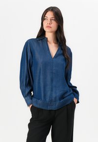 Mörkblå denimskjorta med v-ringning, långa puffärmar och avslappnad passform. Har synliga sömmar och en mjuk textur.