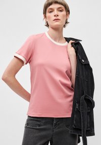 Ροζ βαμβακερό t-shirt με λευκή αντίθεση στον γιακά και στα μανίκια, με απλό σχέδιο. Το μοντέλο φοράει μια μαύρη τζιν ζακέτα περασμένη στον έναν ώμο.