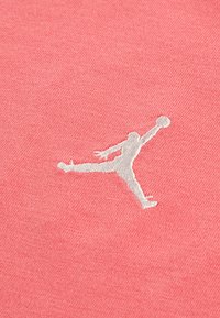 Světle růžová bavlněná látka má bílou vyšívanou logo Jumpman, znázorňující postavu ve vzduchu s basketbalem. Hladká textura.