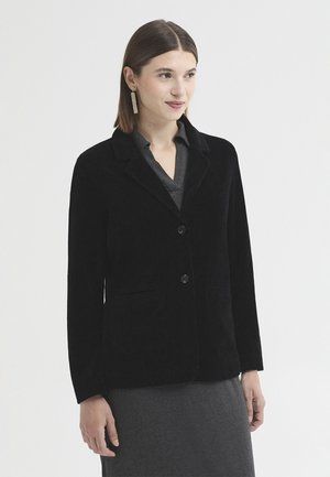 Femme aux cheveux bruns raides portant un long blazer noir sur une robe gris foncé, debout devant un fond clair uni, regardant sur le côté.