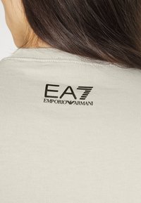 Ljust grå ribbad t-shirt med en svart logotryck på baksidan med texten "EA7 EMPORIO ARMANI". Slät textur och figurnära design.