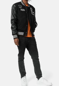 Mężczyzna ubrany w czarną kurtkę typu varsity z naszywkami, pomarańczową koszulkę, czarne dżinsy i białe trampki, stojący na białym tle.