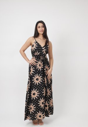 Vrouw staat met één hand op haar heup, draagt een zwarte mouwloze maxi-jurk met beige zonpatronen, tegen een effen witte achtergrond.