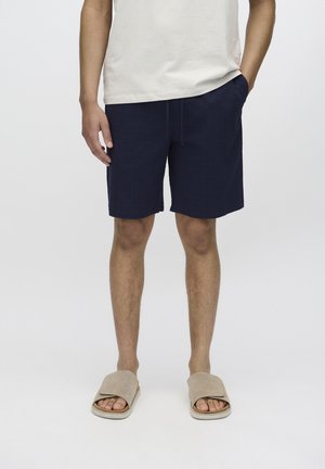 Shorts bleu marine en tissu texturé, avec poches latérales et coupe droite, associés à des sandales beige à enfiler.