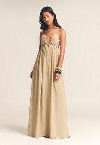 Vestido largo, beige, con escote en V profundo, detalles fruncidos en el busto y tirantes finos. Presenta una falda fluida y cierre con lazo.