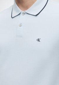 Ανοιχτό μπλε πόλο με δύο κουμπιά και σκούρη λεπτομέρεια στον γιακά, με μικρό σκοτεινό λογότυπο «cK» στο αριστερό στήθος.