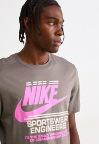 Pilkas medvilnės marškinėliai su dideliu rožiniu Nike logotipu ir tekstu. Trumpomis rankovėmis, apvaliu iškirpimu ir minimalistiniu dizainu su grafikos akcentais.