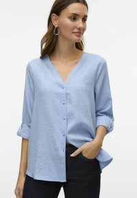 Svijetloplava košulja s dugmadi, izrezana u obliku slova V, s tri četvrtine rukava i mekim materijalom. Uparena s tamnim trapericama, što stvara ležeran stil.